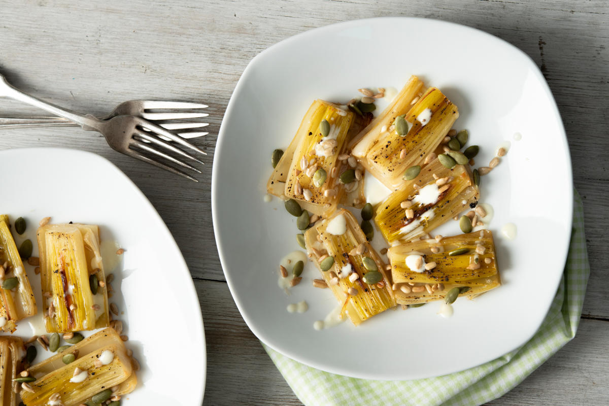 Grilled leeks topped with seeds and a creamy drizzle on white plates, resting on a light wooden table with a checkered napkin and forks.