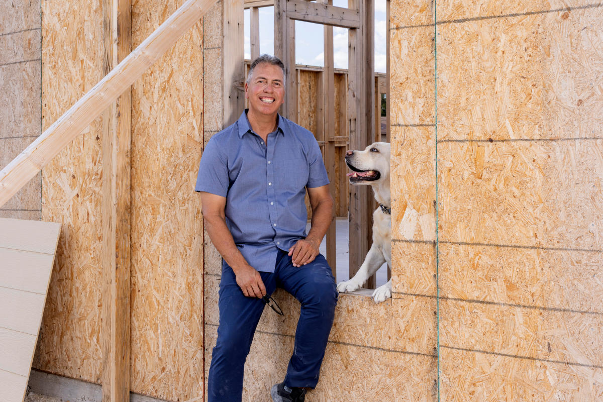 Man smiling at a construction site, wearing a blue shirt and jeans. He leans against an unfinished wooden wall with a happy dog looking out a window.