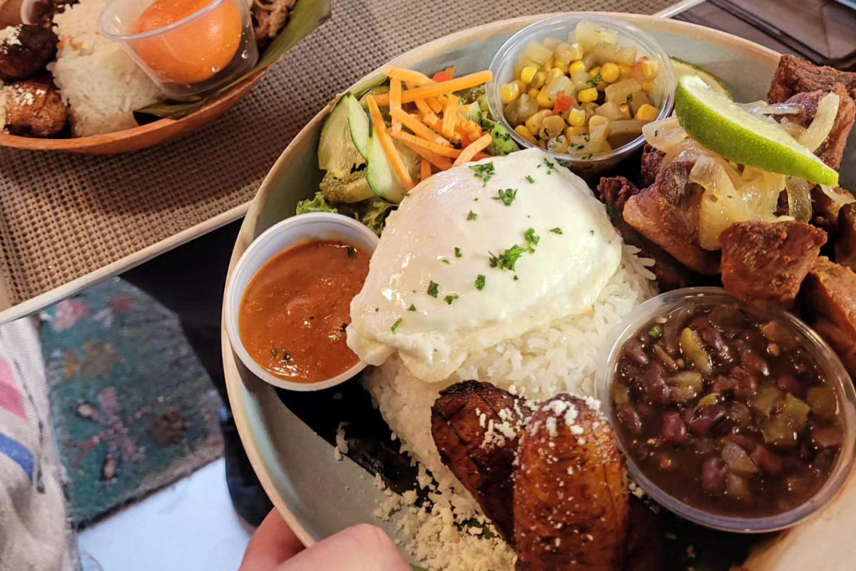 Plate of Colombian food with rice, fried plantains, avocado, beans, corn salad, and a poached egg.