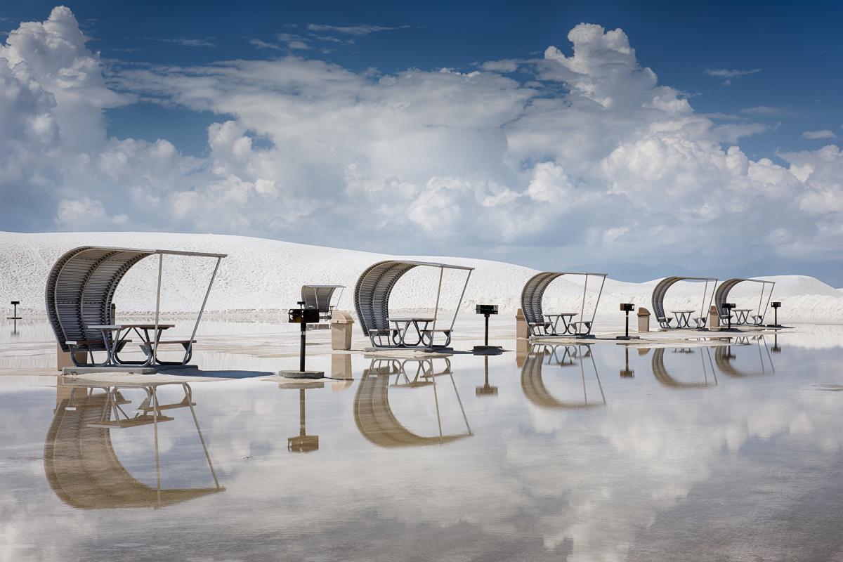Curved picnic shelters and grills reflected on a wet surface in a bright white desert.