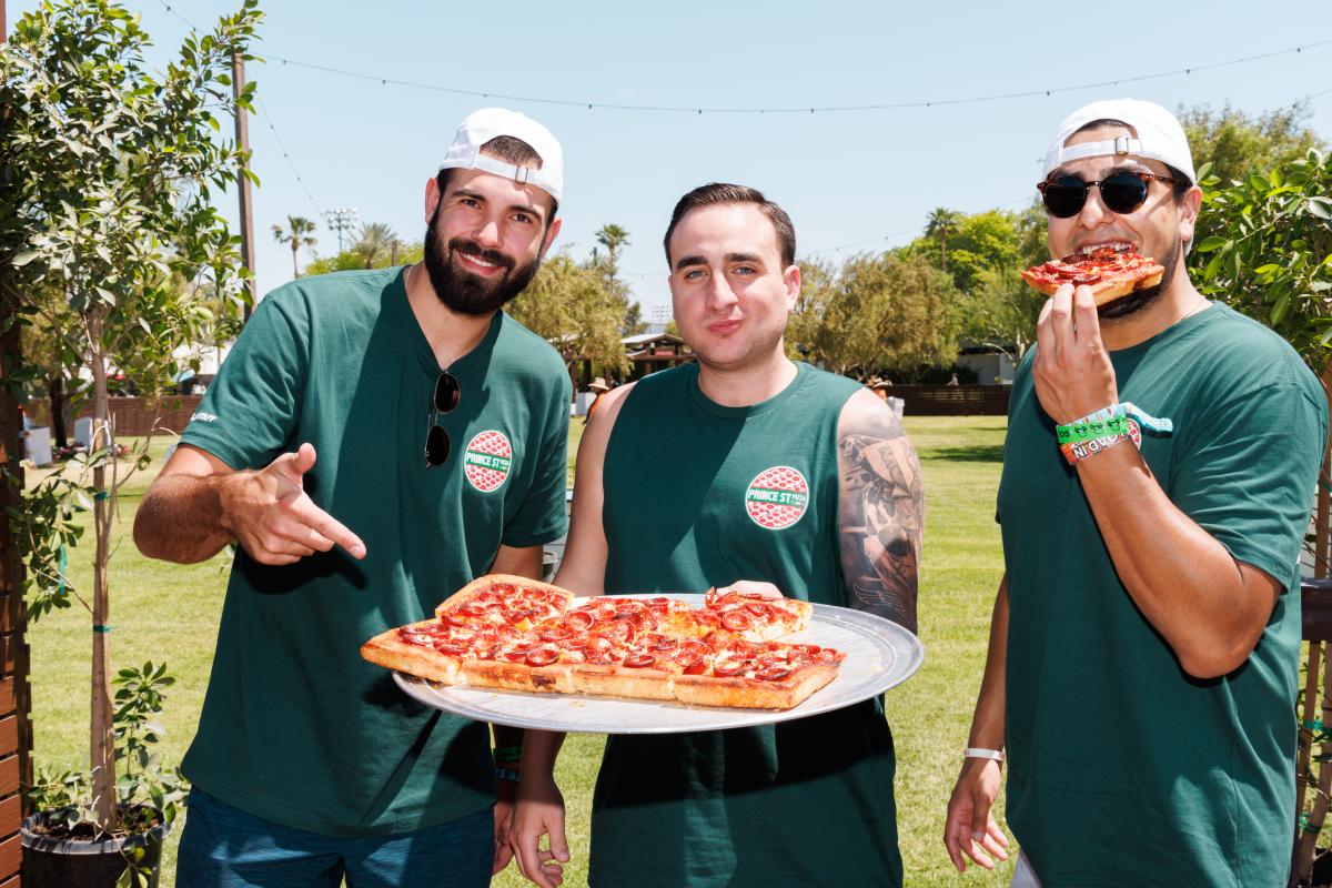 Three men hold up a pepperoni pizza from Prince St. Pizza.