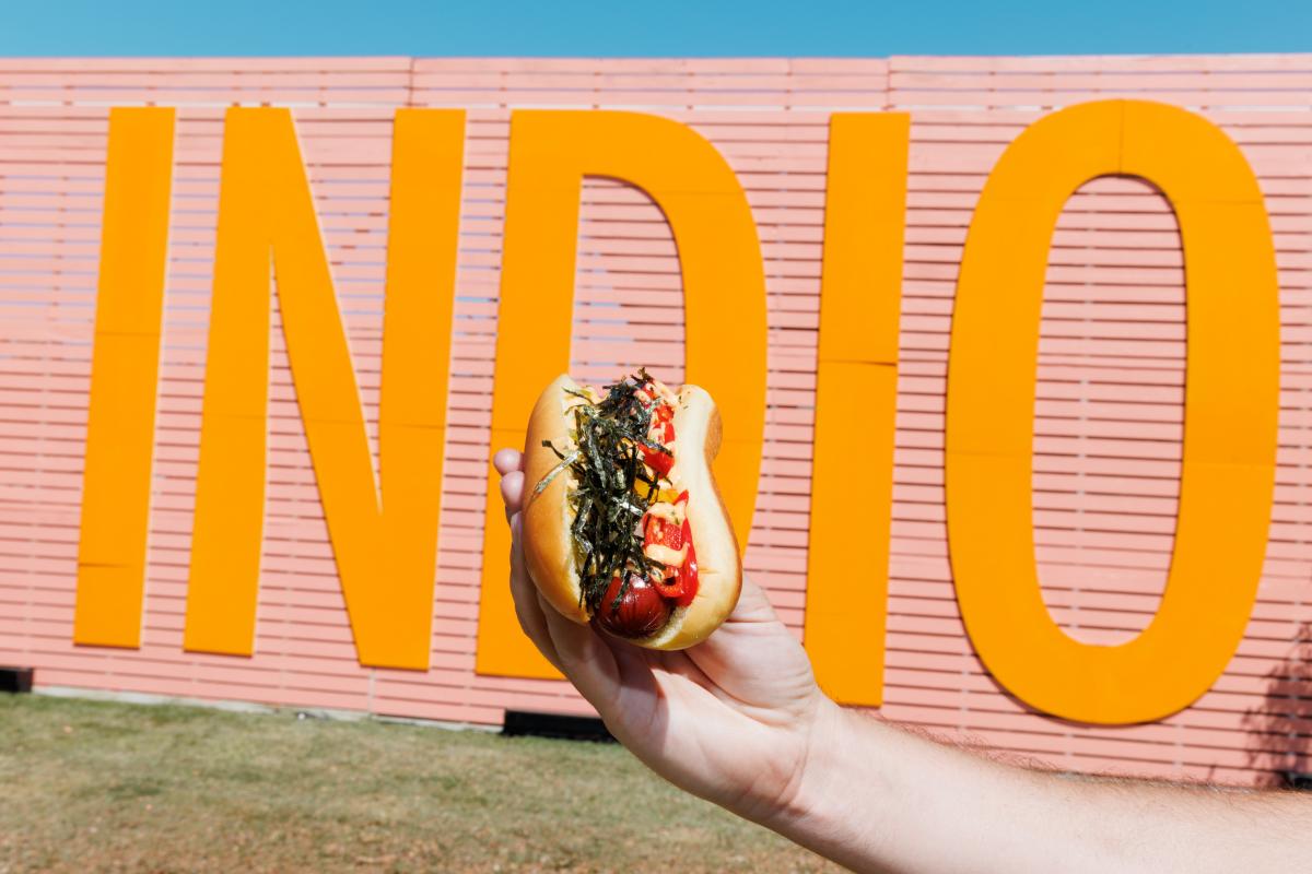 A person holds up a gourmet hot dog in front of the Indio Central Market sign at Coachella Valley Music and Arts Festival.
