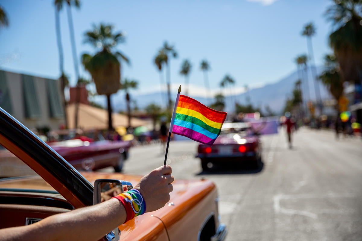 Pride flag waving outside of car in downtown Palm Springs with mountains and palm tree