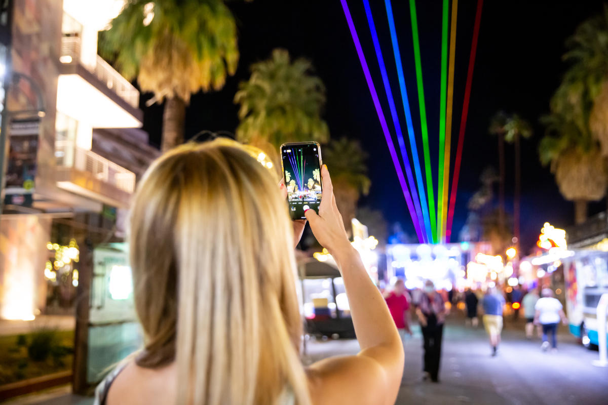 Blonde woman taking a photo of rainbow lasers with her phone during a Pride festival in Palm Springs at night