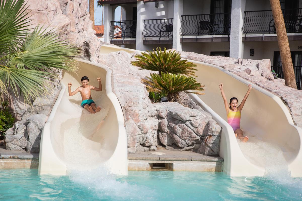 two children on waterslide splashing into water palm trees vacation beautiful clear blue pool