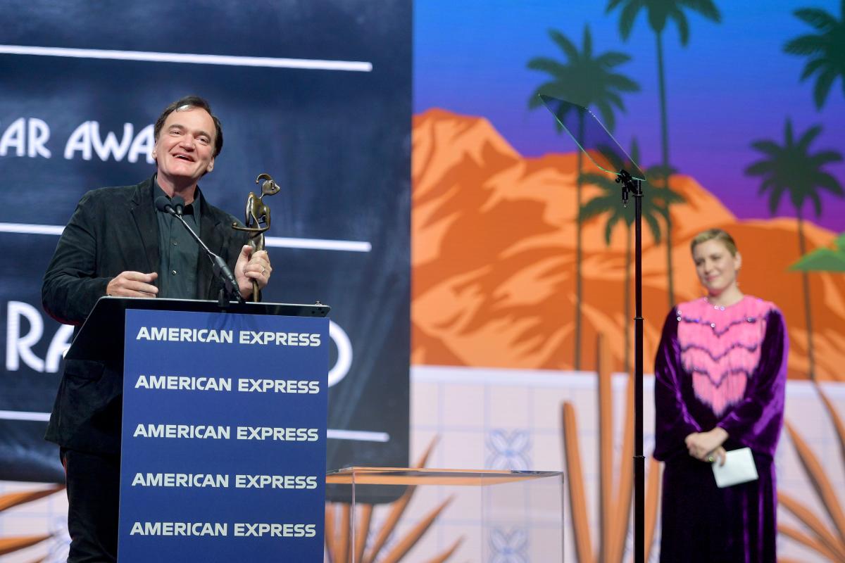 Quentin Tarantino accepts an award at the 2020 Palm Springs International Film Festival Film Awards Gala with Greta Gerwig in the background.