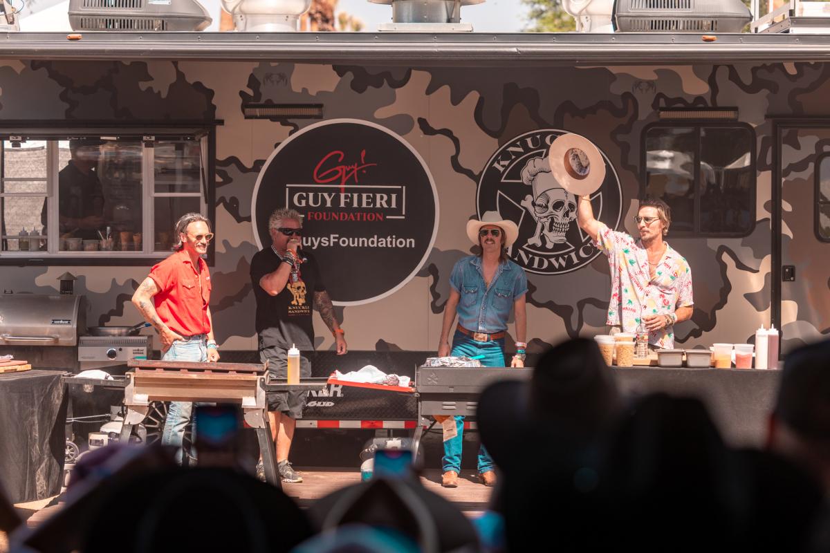 Guy Fieri and his friends in front of an RV leading a cooking demonstration for fans.