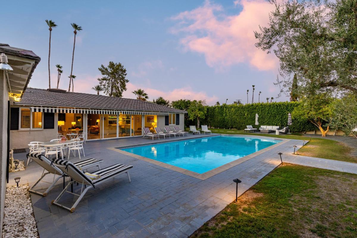 A serene twilight view of a backyard with a swimming pool, lounge chairs, outdoor seating, and palm trees against a dusky sky.
