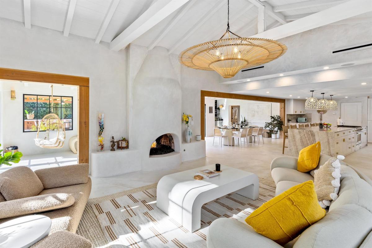 A spacious modern living room with white walls, a vaulted ceiling with exposed beams, and large windows. The room features a seating area with plush sofas and a unique white coffee table, a hanging rattan chair, an open fireplace, and decorative lighting. In the background is an open-plan dining area and a kitchen with white cabinetry and an island.