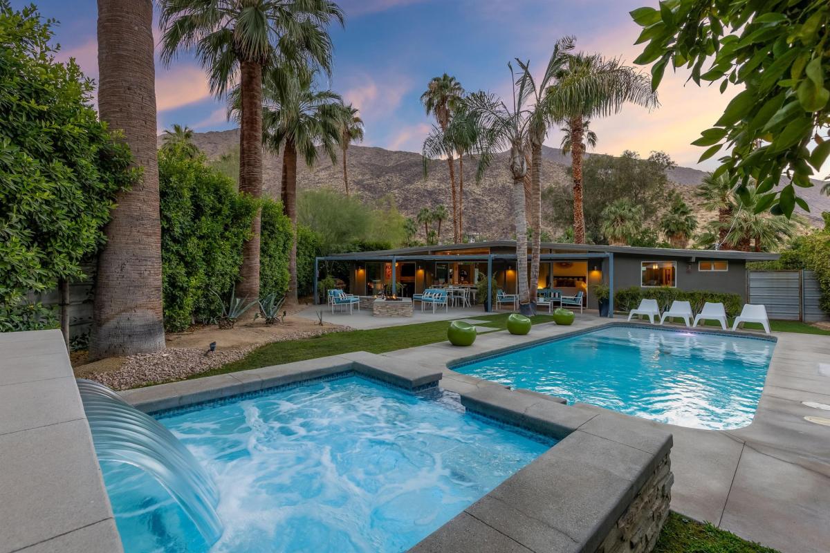Image of an elegant backyard with a swimming pool and a modern house against a backdrop of mountain and palm trees during twilight.