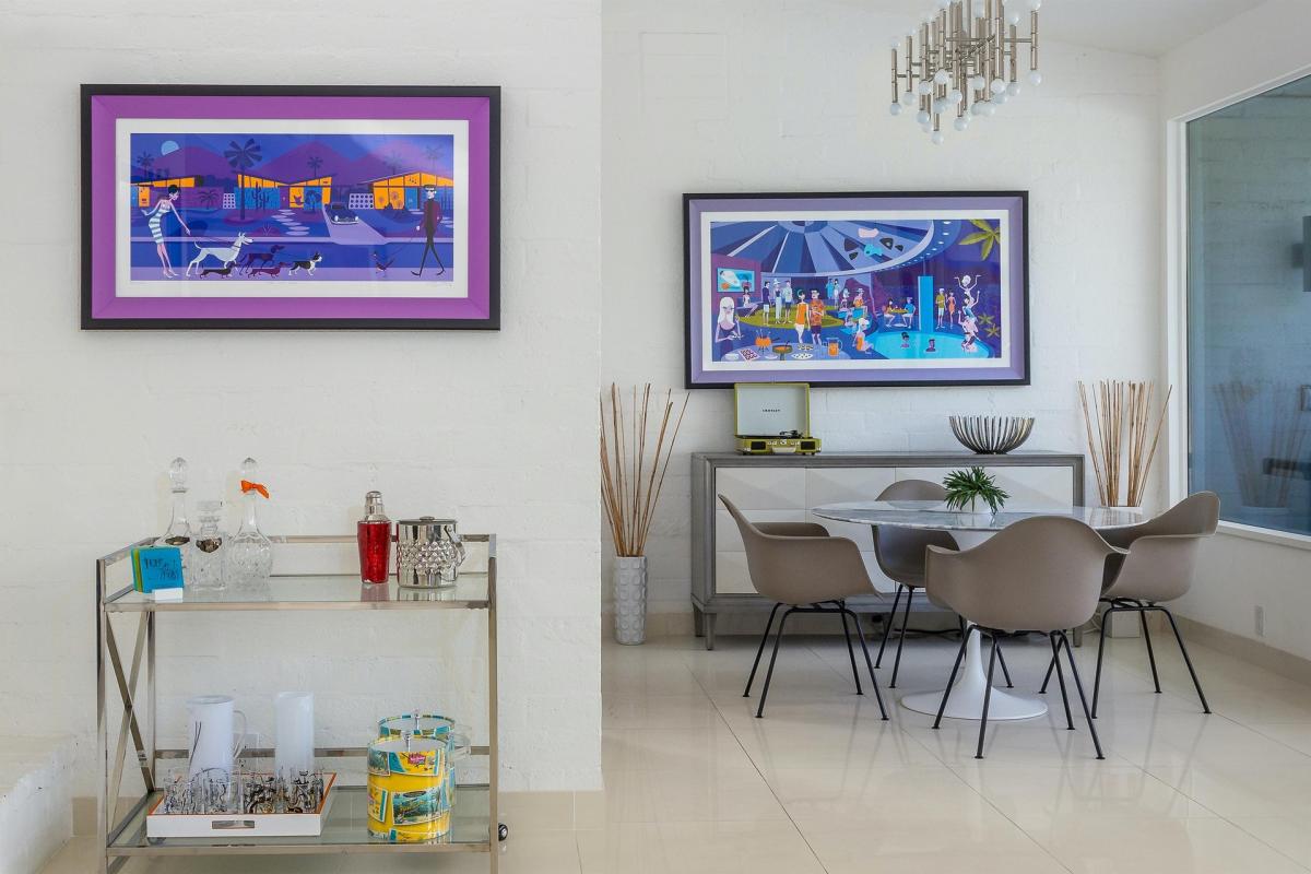 A contemporary dining room with modern artwork on the walls, featuring a glass bar cart and a dining table with grey chairs.