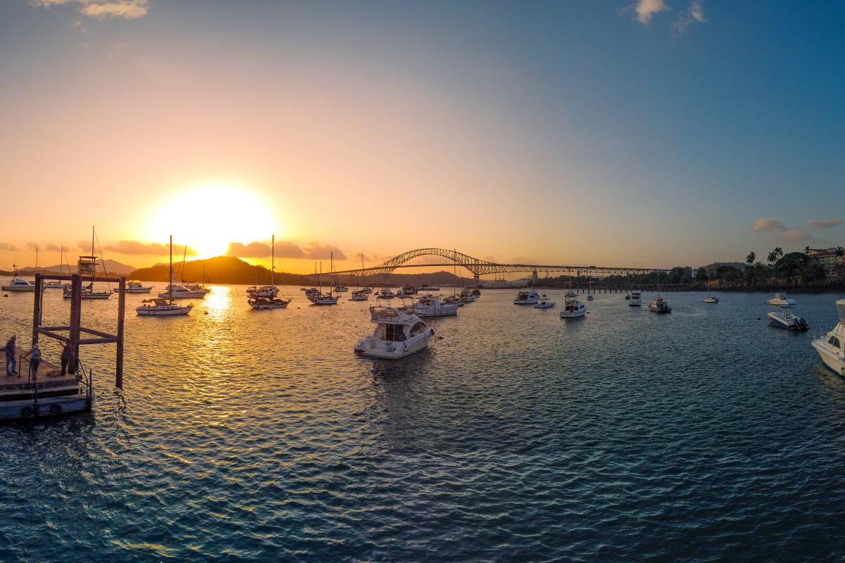 Beautiful sunset over a harbor. The sun is setting behind a bridge of the Americas in the distance, casting a golden light on the water. There are many boats anchored in the harbor, and people can be seen walking along the docks.