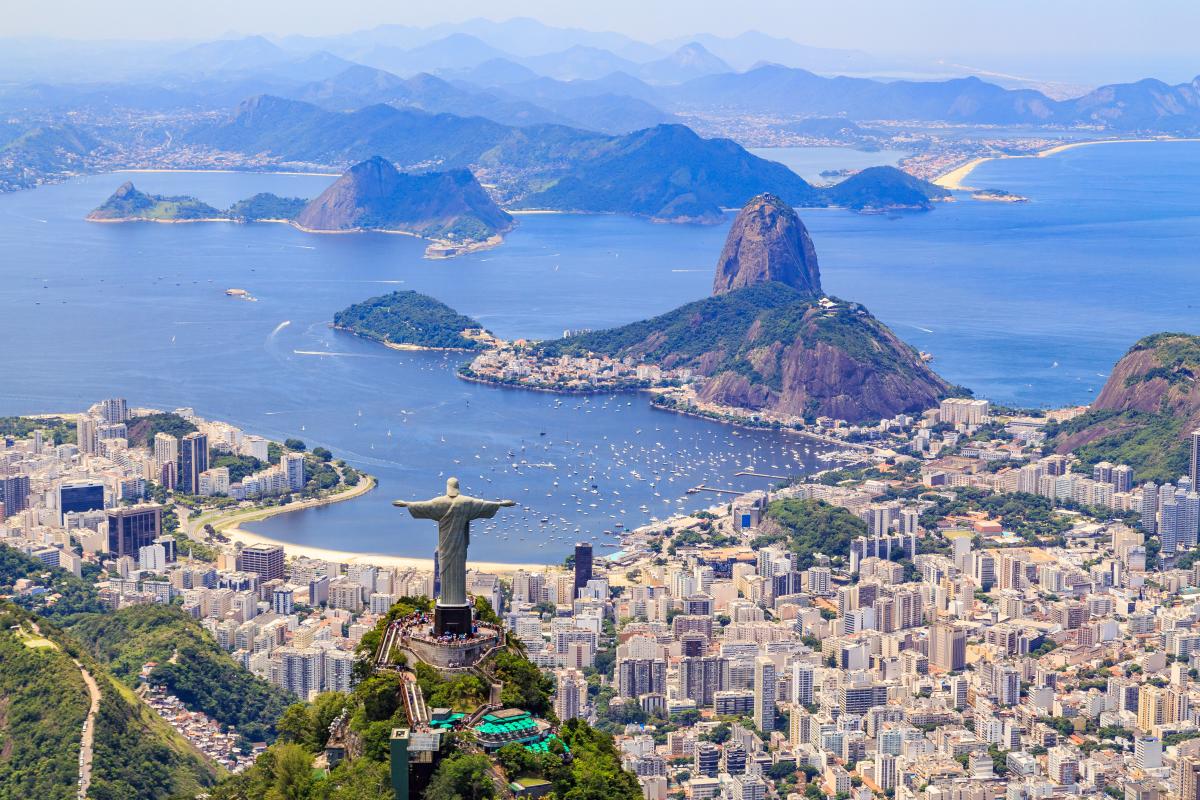 An aerial view of Rio de Janeiro, Brazil, featuring the iconic Christ the Redeemer  statue atop Corcovado Mountain. The city is spread out below, with Guanabara Bay  and Sugarloaf Mountain visible in the distance.