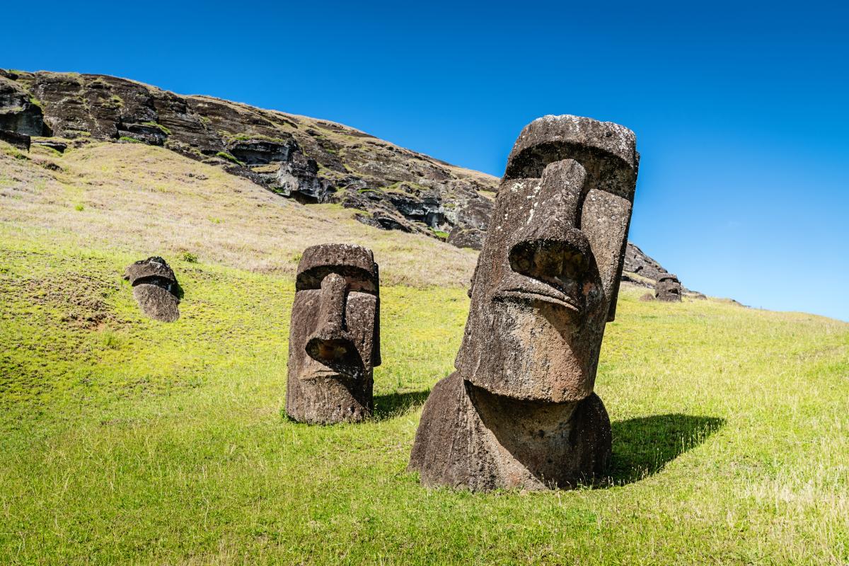 Several large, stone Moai statues standing in a field on Easter Island. The statues have elongated ears and stoic expressions, with a backdrop of rolling hills and a blue sky.