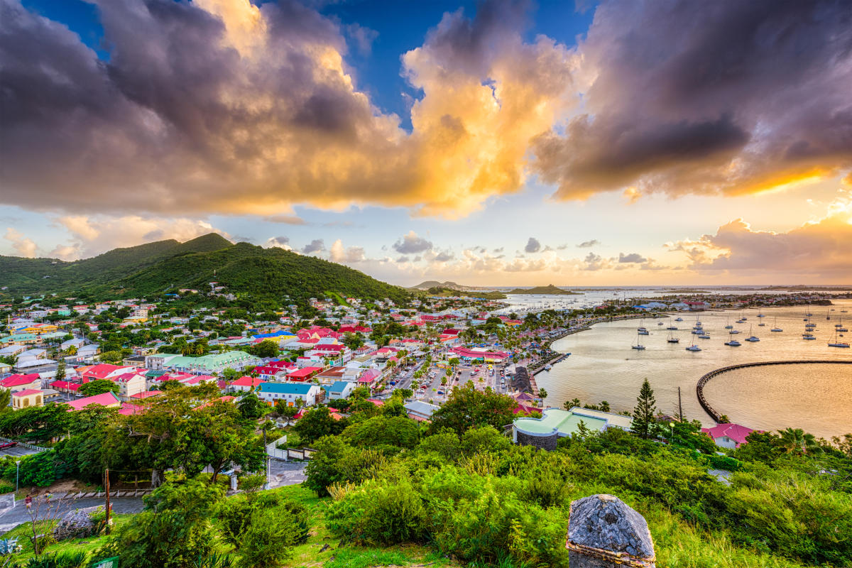 A scenic aerial view of Marigot, Saint Martin, at sunset. The town is nestled between hills,  with colorful buildings and a vibrant waterfront. Boats are anchored in the harbor, and dramatic clouds fill the sky.