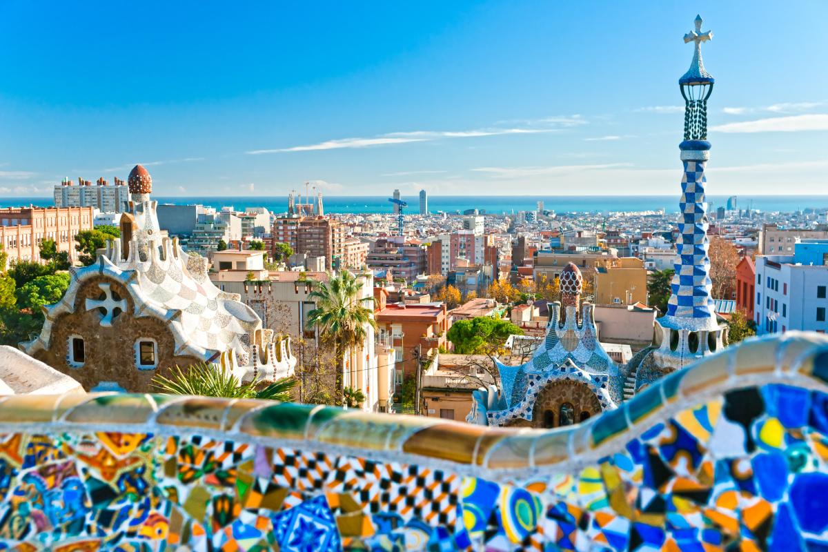 A panoramic view of Barcelona, Spain, from Park Güell. The iconic mosaic bench and colorful buildings are visible, with the city skyline and the Mediterranean Sea in the background.