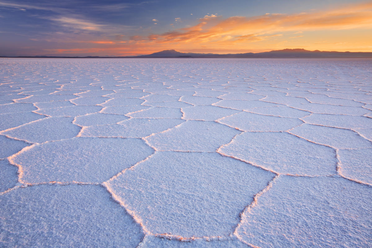 A vast, flat salt pan with hexagonal patterns formed by salt crystals. The sky is a vibrant orange and pink at sunset, and mountains can be seen in the distance.