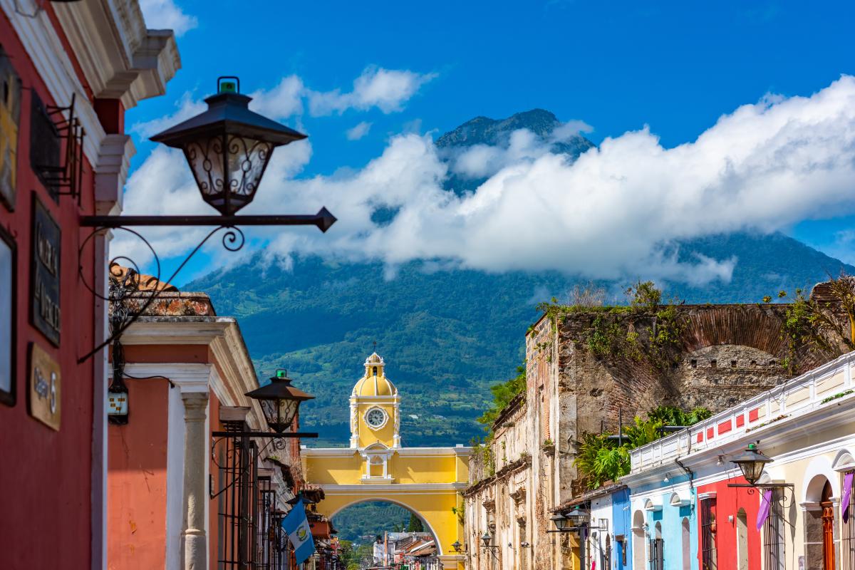 A colorful street in Antigua, Guatemala, with a yellow archway and a clock tower.  The street is lined with brightly colored buildings, and a volcano with a cloud-covered  peak rises in the background.