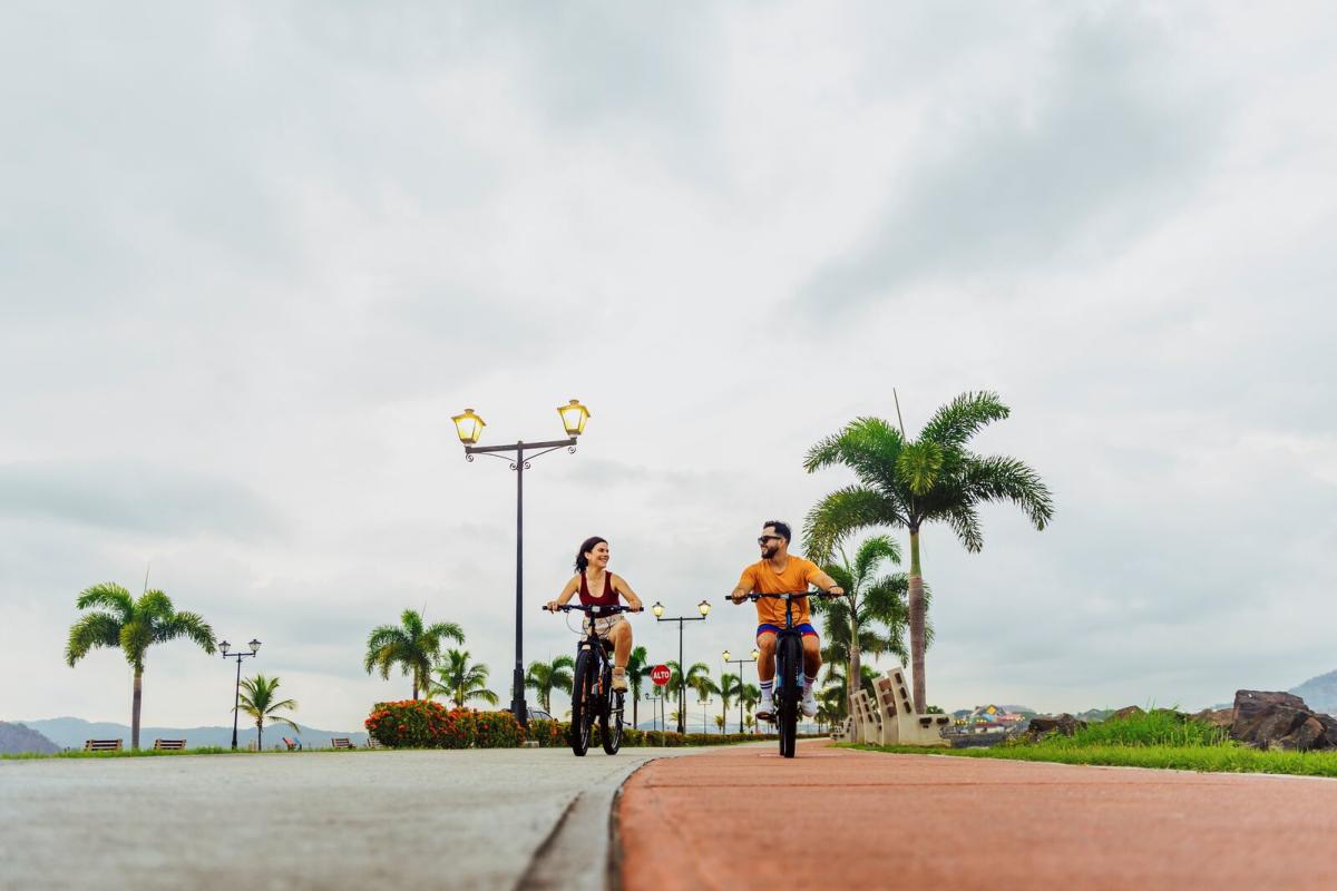 A happy couple rides bicycles on the Amador Causeway, with the palm-lined road visible, the sea on the side, on a sunny day.