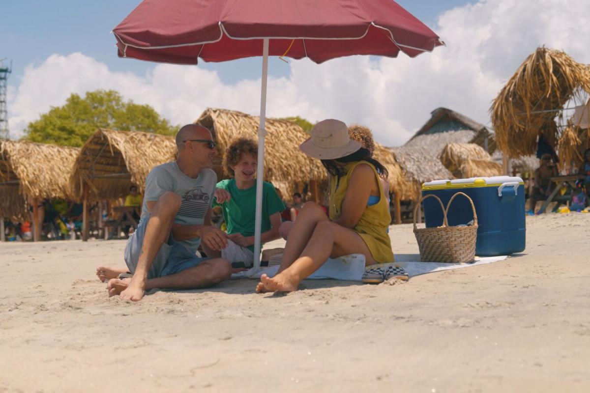 Uma família senta-se sob um guarda-sol vermelho, sorrindo e relaxando em uma praia de areia em Panamá, pontilhada de cabanas de palha, sob um céu parcialmente nublado.
