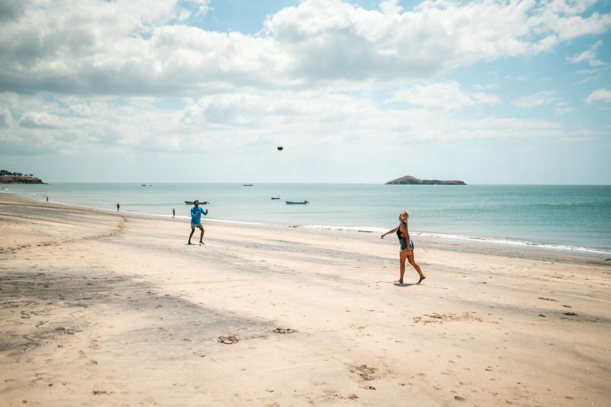 Una escena de playa con dos personas jugando en la arena cerca del agua, con una persona de azul lanzando un objeto hacia otra persona con un bañador oscuro. Al fondo, hay botes en el agua y una isla a la distancia, bajo un cielo parcialmente nublado.