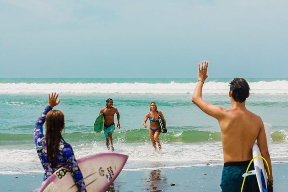 Dois surfistas de fato de banho correm das ondas do oceano em direção a amigos que seguram pranchas de surf e acenam numa praia ensolarada em Panamá, evocando alegria e camaradagem.