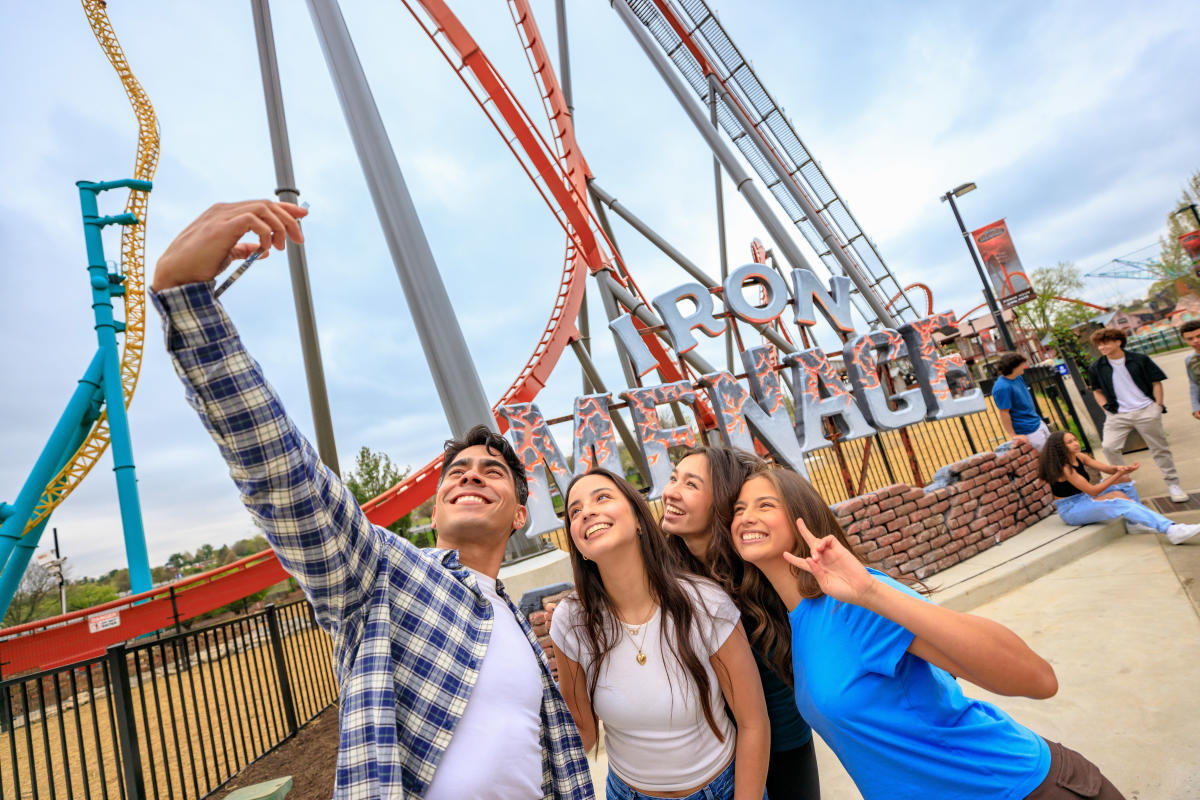 A group of people take a selfie in front of a towering roller coaster with looping red and yellow tracks at an amusement park in Pennsylvania.