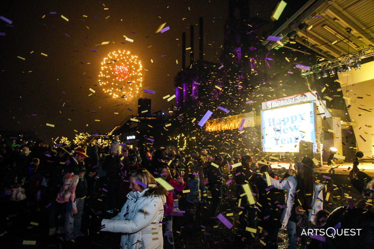 People cheering with purple and yellow confetti in front of a stage with fireworks going off