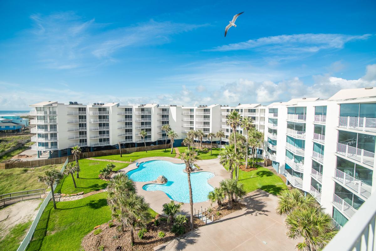 Aerial photo of a beautiful white condominium complex with palm trees, a pool, and green grass.