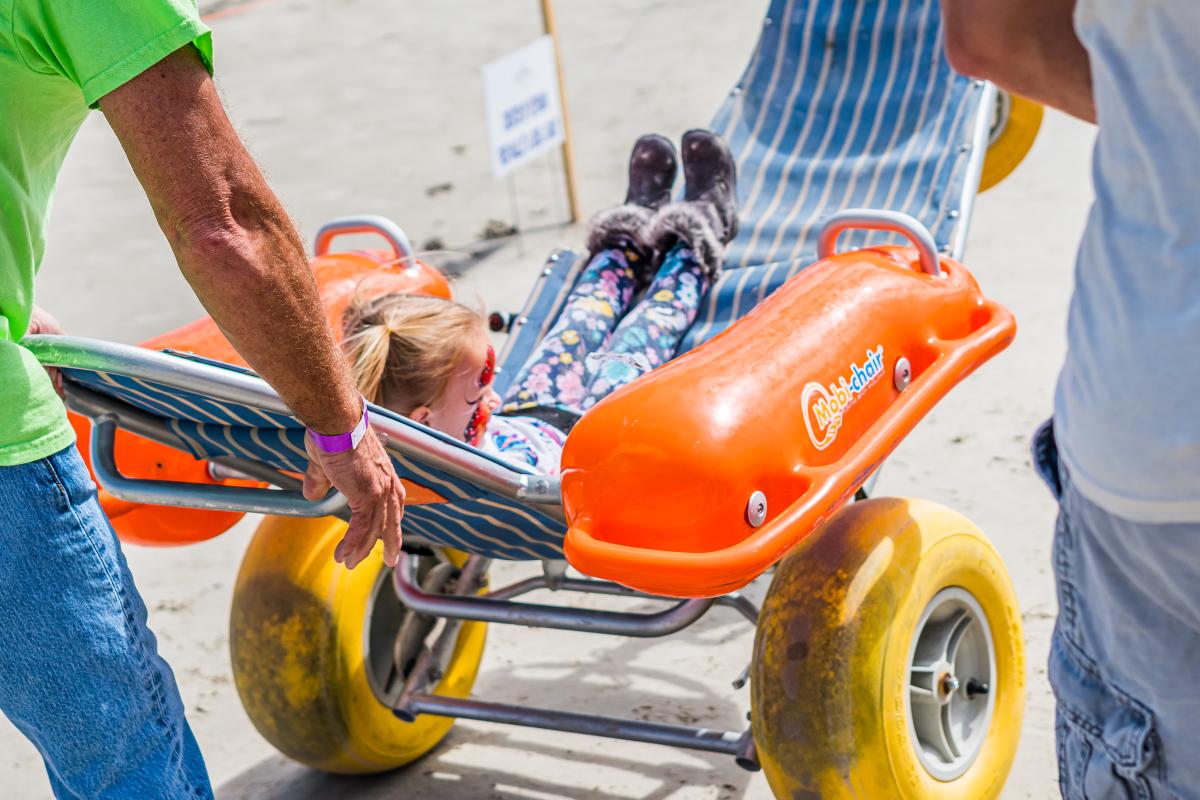 A young girl is pushed on the beach in a large-wheeled wheelchair