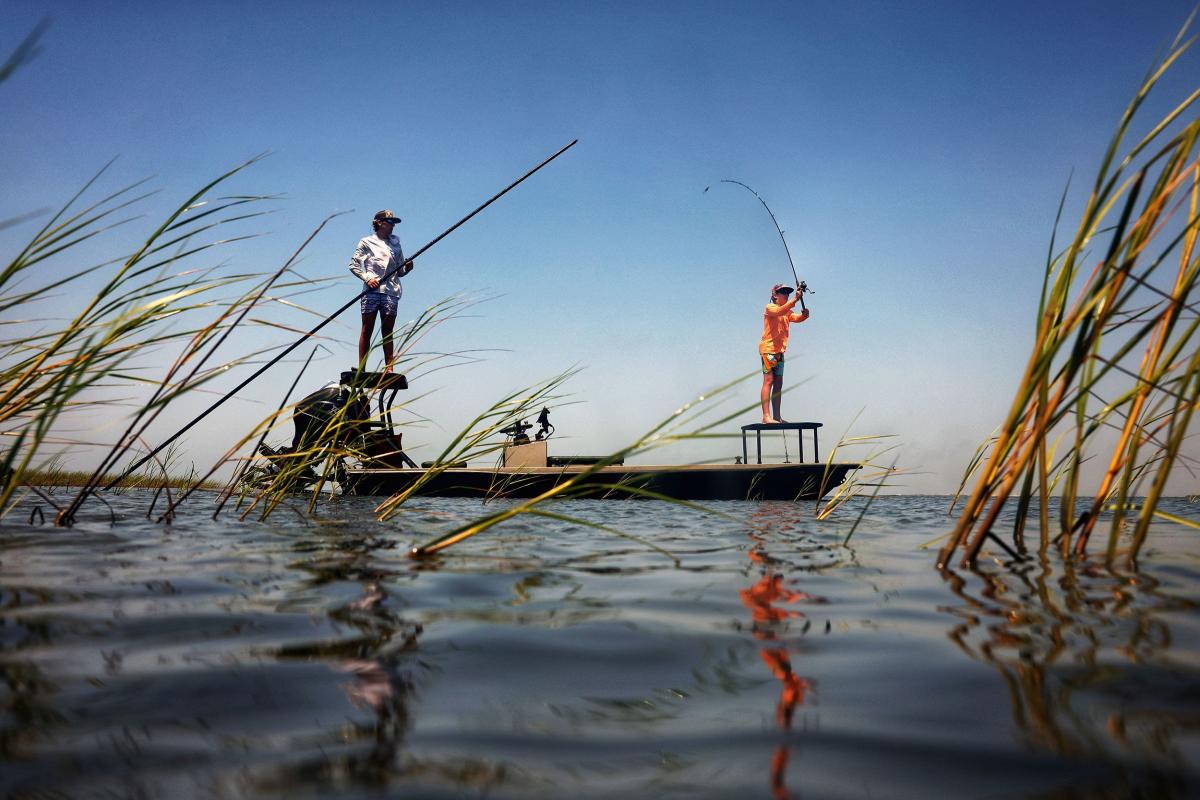 Two fly fishermen on a small boat in a calm marsh at sunset, surrounded by tall seagrass.