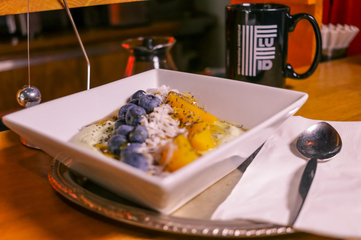 A square white bowl with fruit and cream sits in front of a black coffee mug