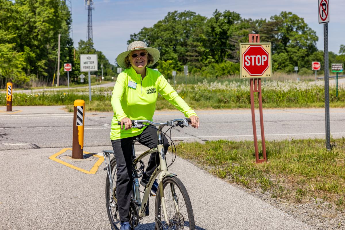 A woman wearing a wide-brimmed hat and green long sleeve shirt is riding a bike on a paved trail.