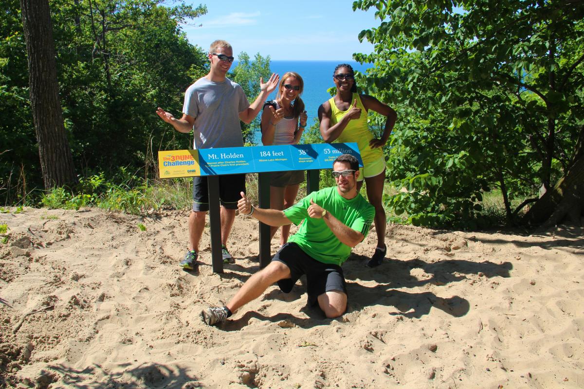 A group of four friends poses at the summit of Mt. Holden, a sand dune overlooking Lake Michigan. They stand around a blue and yellow sign marking the elevation of 184 feet. One person in a bright green shirt kneels in the sand, pointing at the sign with enthusiasm, while the others strike playful poses. The background features lush green trees and a clear blue lake under a bright sky.