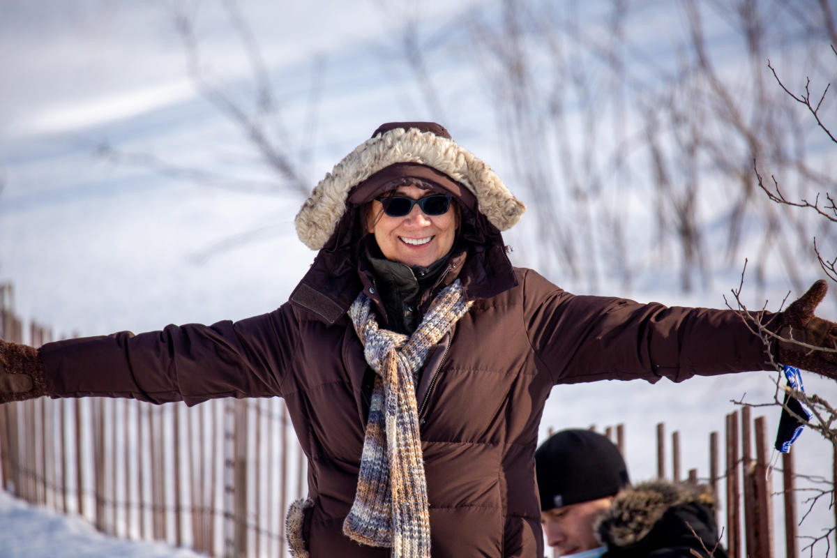 A woman bundled up in winter gear smiles and holds are arms out