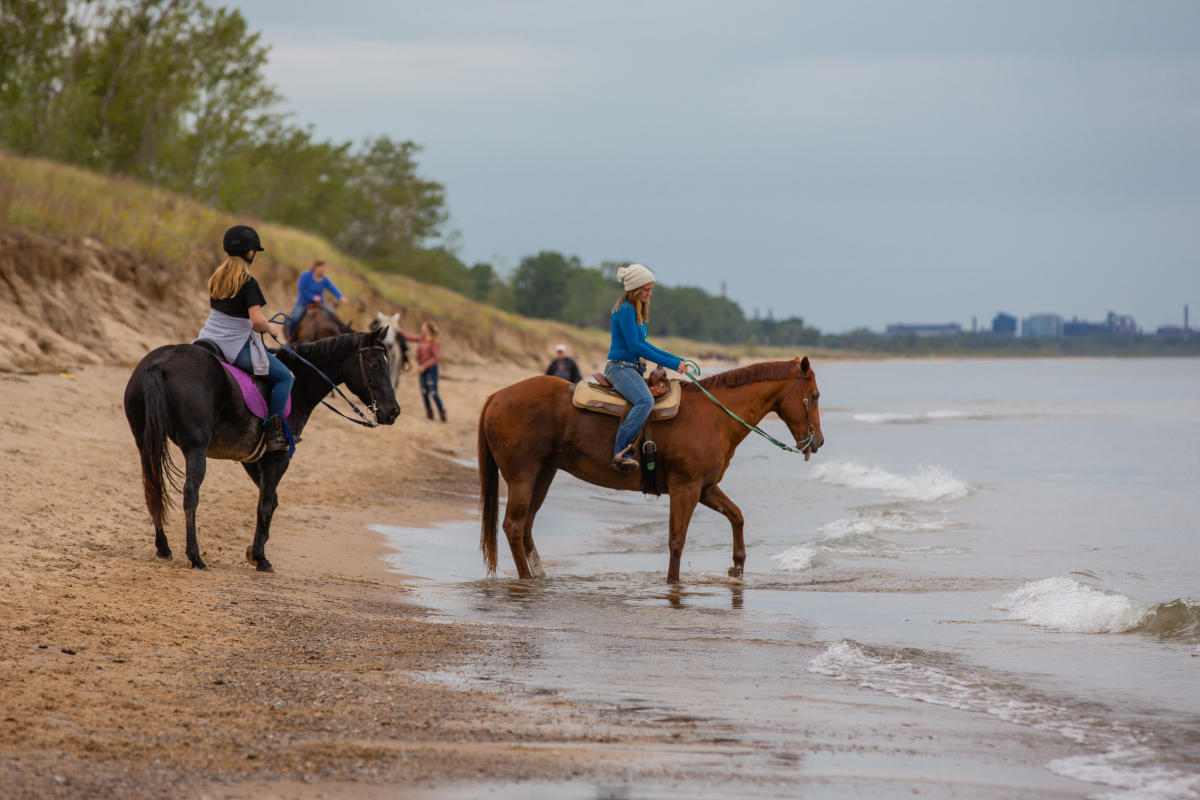 People sit atop horses on the beach at the edge of Lake Michigan. Sand dunes and trees are seen in the background. Industry is seen on the far horizon.