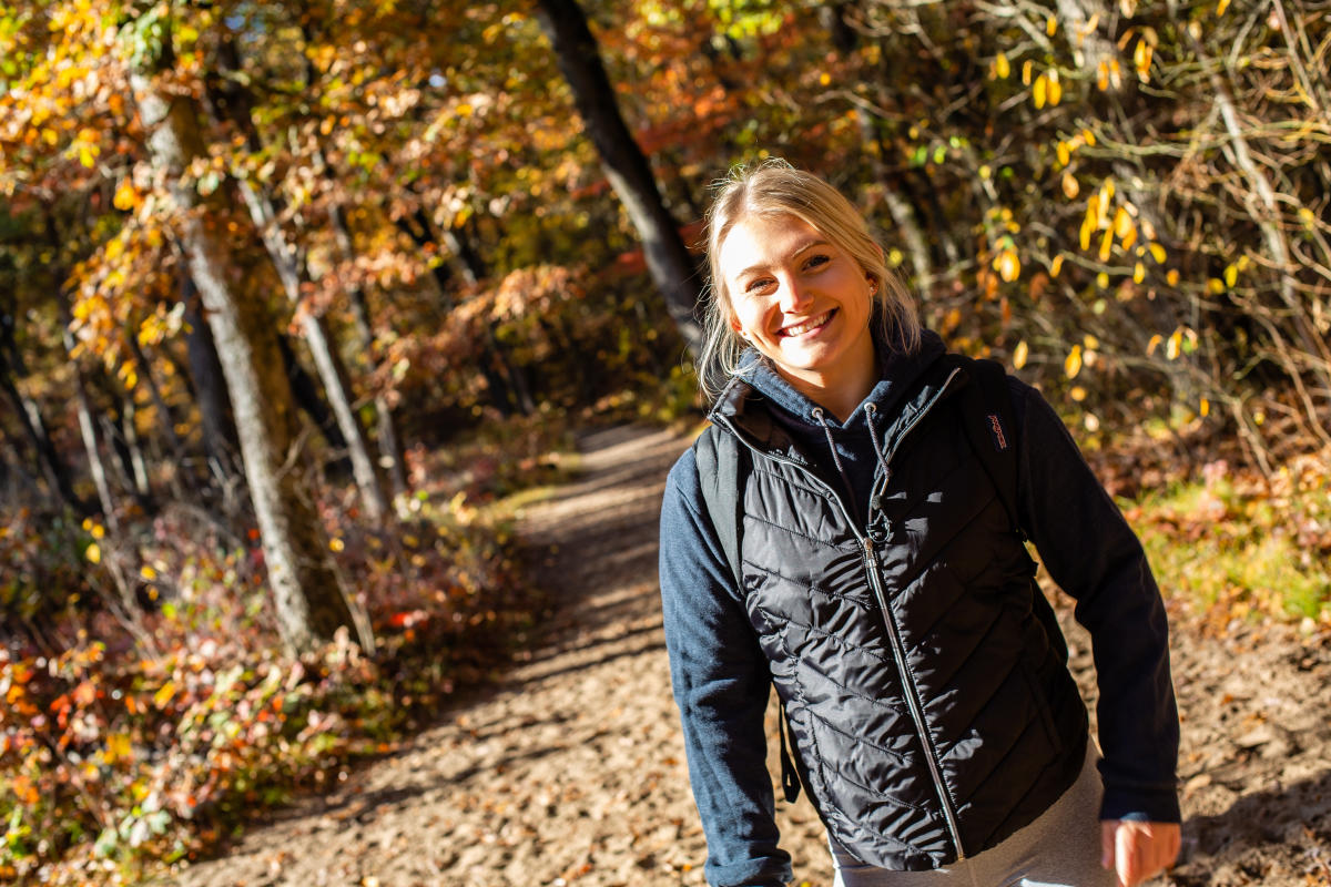 A young blonde woman wearing a dark fall jacket stands in front of a sandy trail surrounded by fall colors.