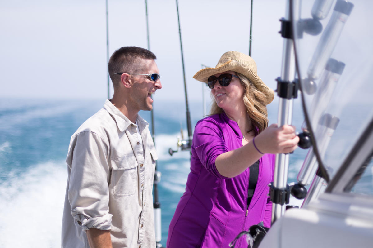 A cheerful moment between two people on a fishing boat, with the open water behind them. The woman, wearing a cowboy-style hat and sunglasses, holds onto the boat's railing, while the man, dressed in a button-up shirt and sunglasses, laughs. The fishing rods in the background hint at a day of adventure on the water.