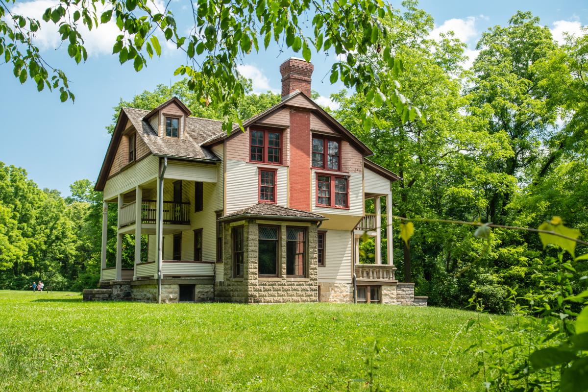 A historical home, tan and peach in color, sits in a field surrounded by trees.