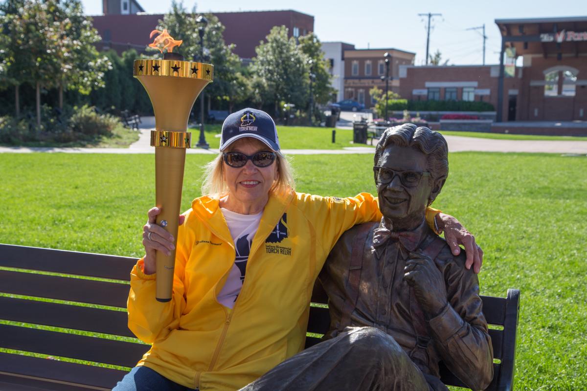 A person holds the Indiana Bicentennial torch while sitting on a bench next to a statue