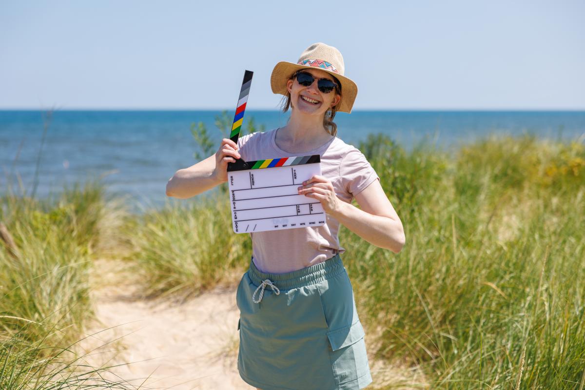 A smiling woman stands on a sandy path leading to Lake Michigan, surrounded by tall green beach grass. She wears a straw hat with a colorful band, sunglasses, a light pink t-shirt, and a sage green skirt with pockets. She holds a film clapperboard with multicolored stripes at the top, raising the clapper with one hand while gripping the board with the other. The lake and a clear blue sky stretch out in the background.