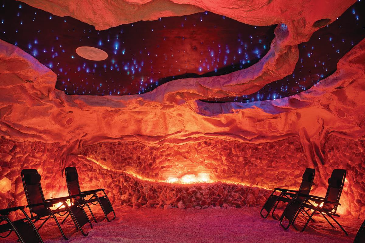 A salt cave relaxation room with textured, reddish-orange salt walls and a floor covered in fine salt crystals. The ceiling mimics a night sky, with twinkling blue star-like lights and a circular moon design. Several black reclining lounge chairs are arranged in a semi-circle, facing the softly glowing salt wall. The room has a warm, ambient glow.
