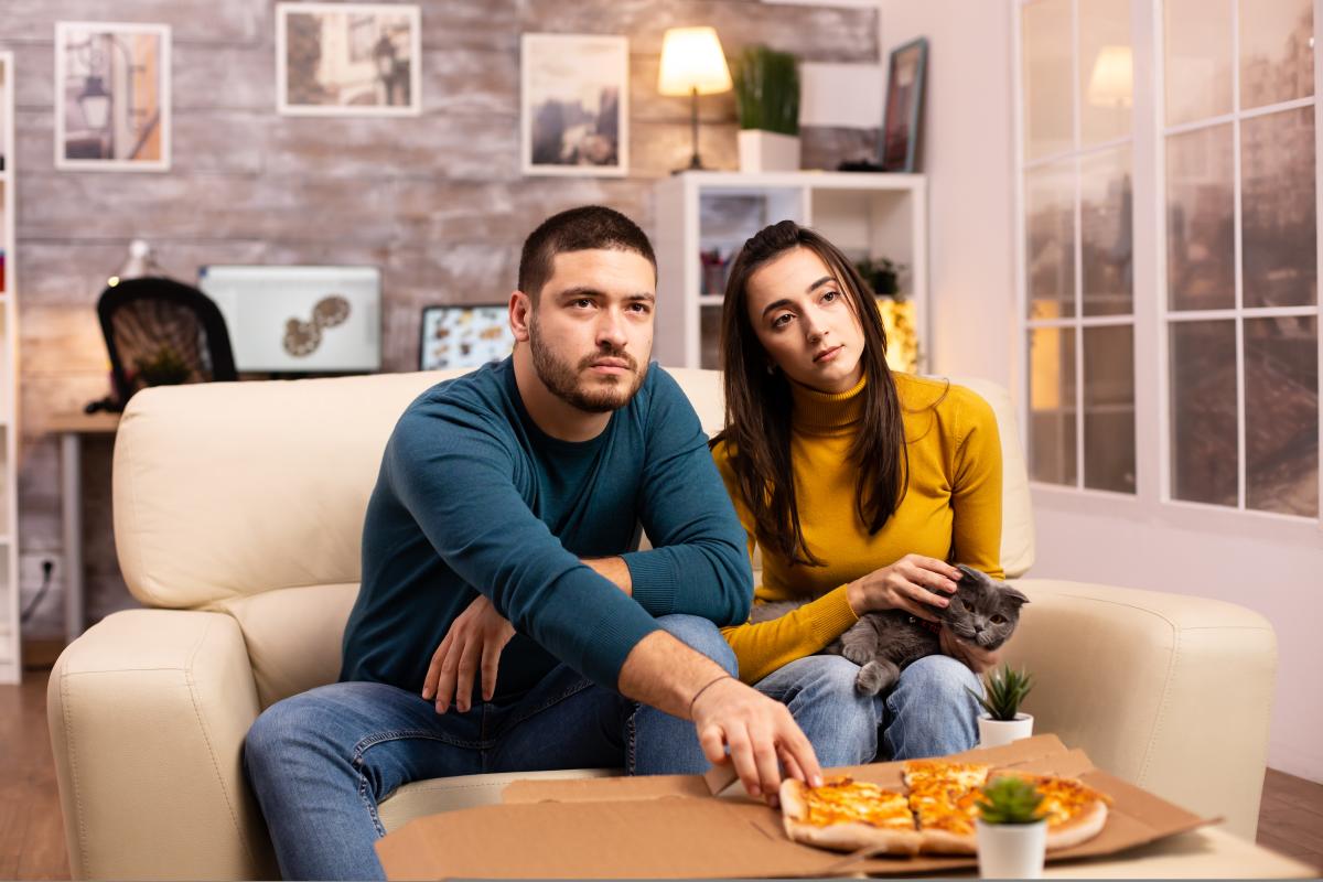 A man and women sit on a couch, looking bored. A pizza is in front of them.