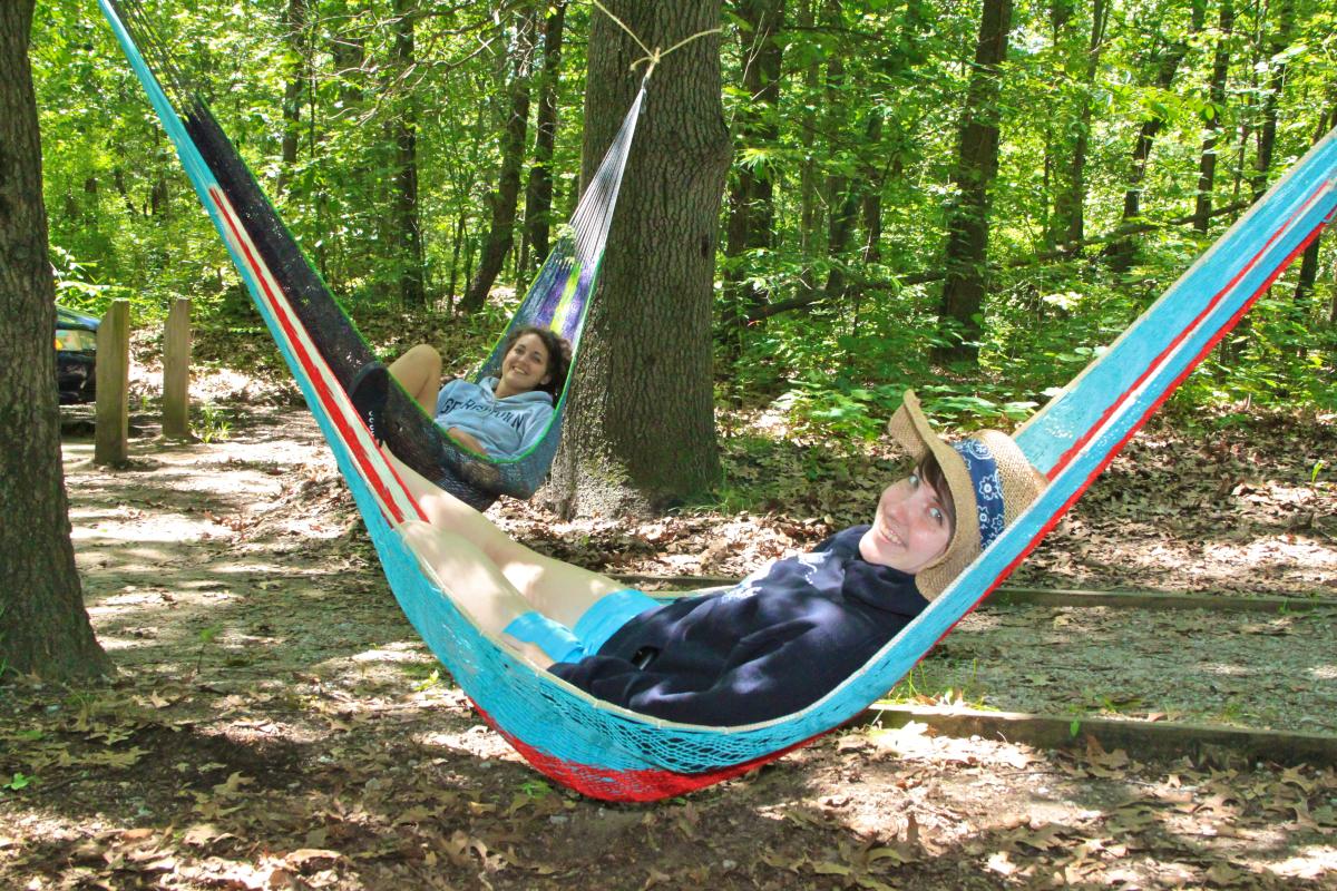 A pair of friends enjoying a forested campsite, lying in hammocks strung between trees. The person closest to the camera, dressed in a hoodie, shorts, and a straw hat, smiles warmly. The second person, wearing a sweatshirt, is reclined in a hammock further back, also smiling. Sunlight dapples the leafy forest floor.