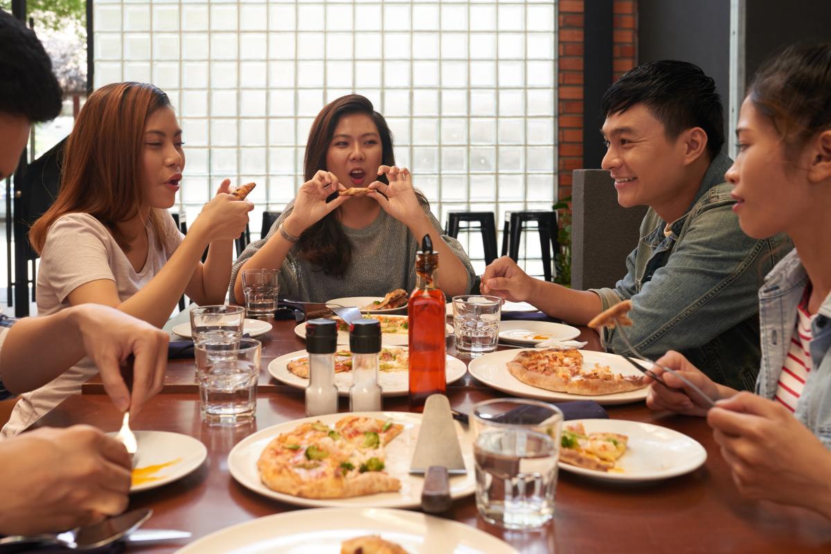 A group of people sit at a restaurant table, chatting and eating pizza