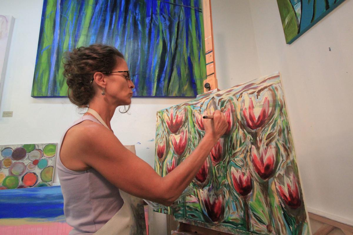 A woman in a tank top and apron paints flowers on a canvas. Other paintings hang on a white wall behind her.