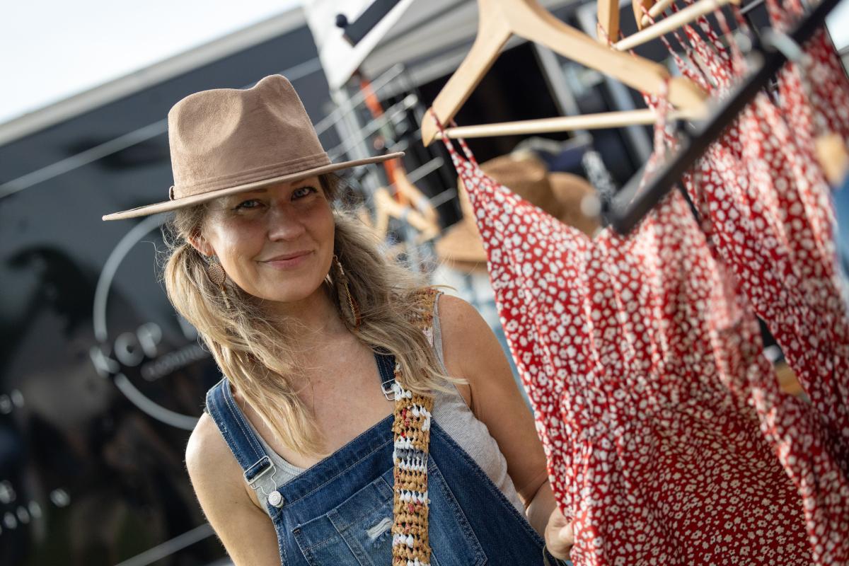 A woman wearing a hat and overalls stands next to clothing on hangers.