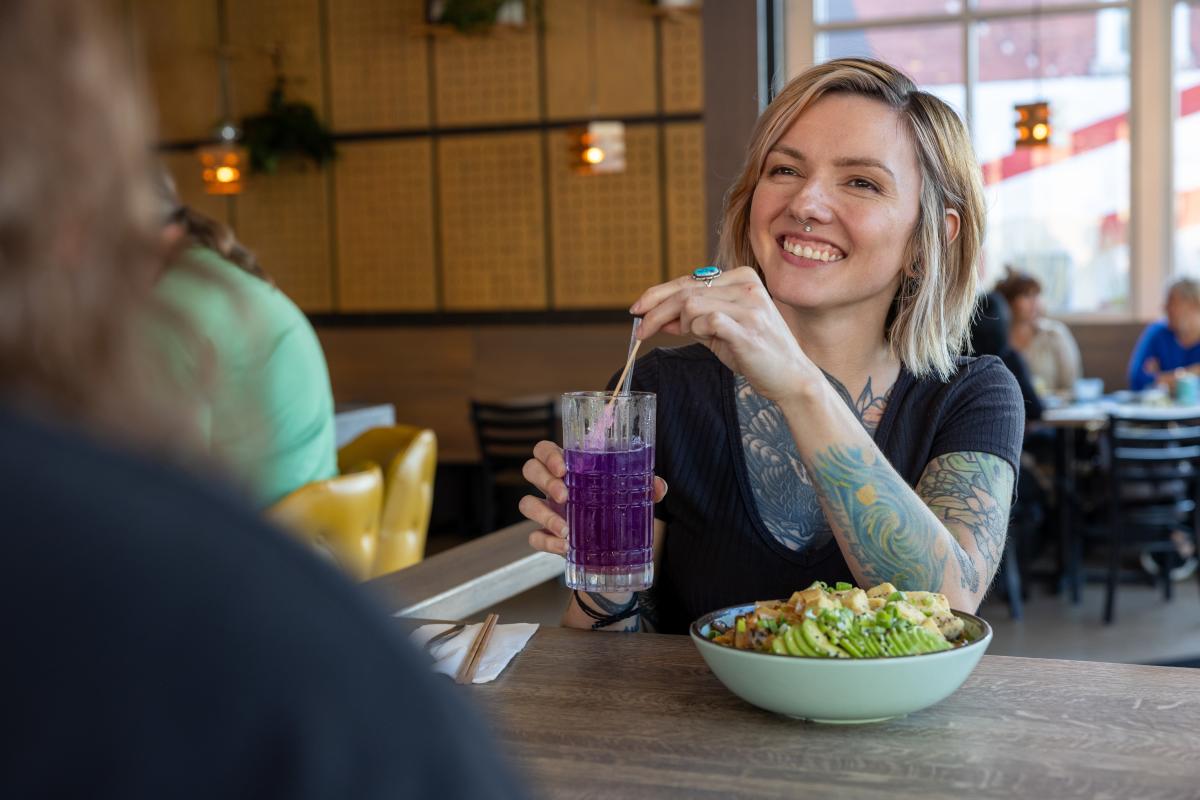 A woman with blonde hair and tattoos holds a purple drink while talking to someone unseen on the other side of the table.