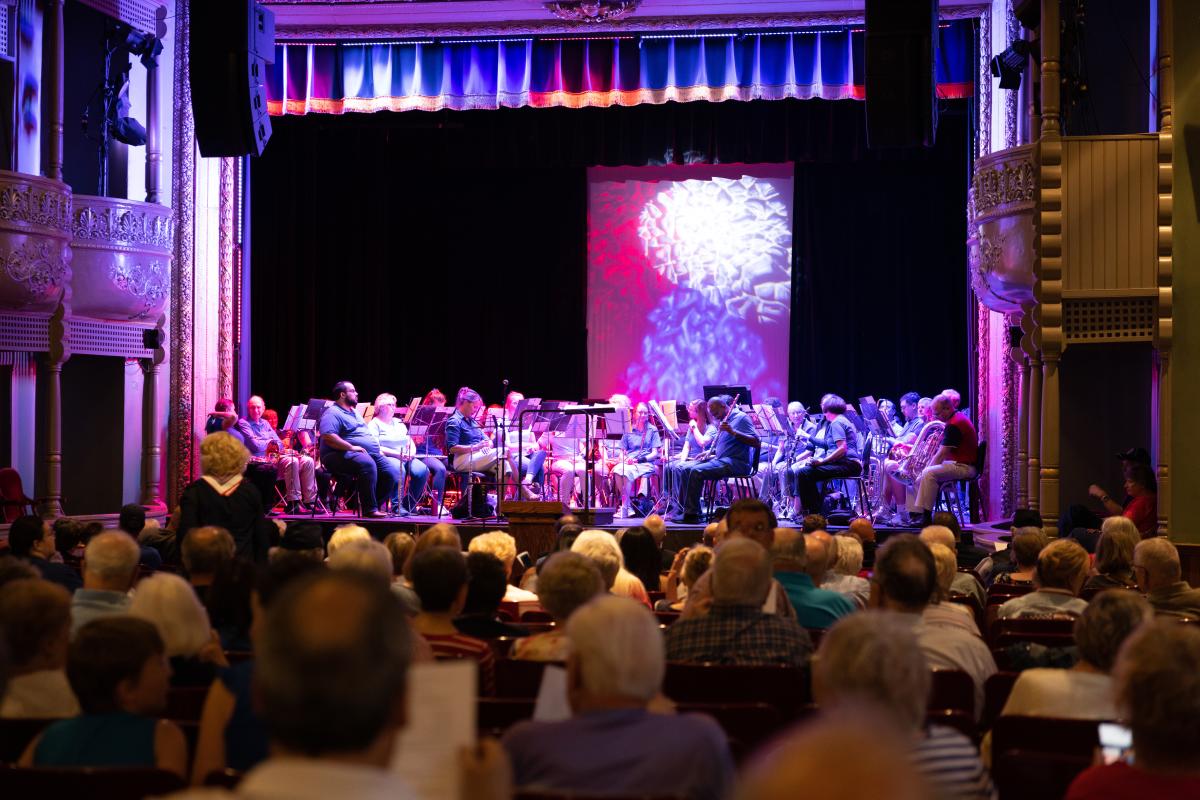 An orchestra plays on stage as the audience watches. The stage is bathed in purple and pink lighting.