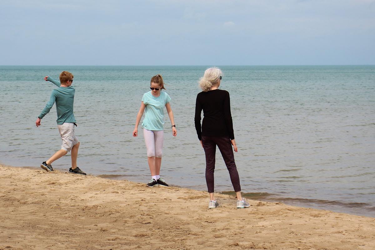 A woman with gray hair and two teens skip rocks on the beach shore.